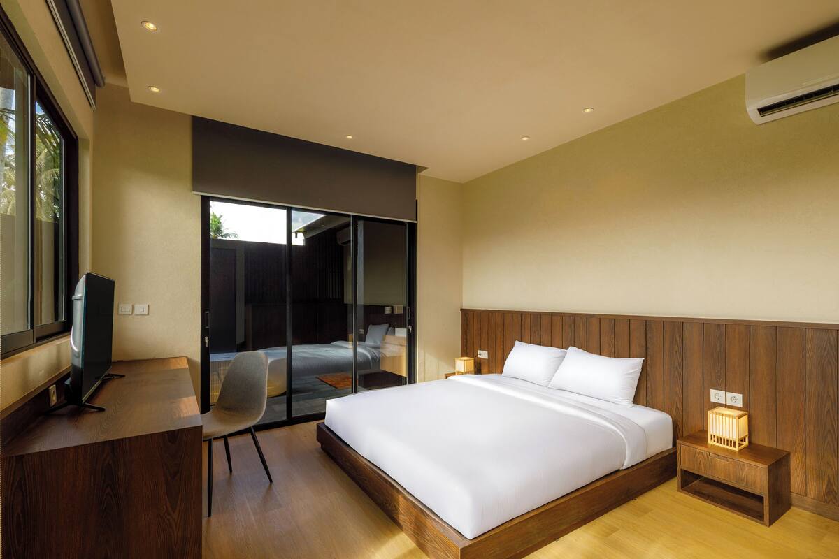 A modern bedroom with a large bed, a desk, a chair, and a sliding glass door leading to a balcony.