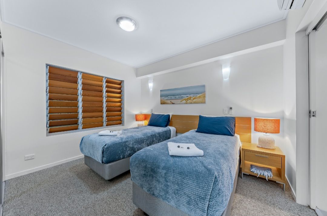 Quarto de hotel com duas camas, janela com persianas, mesa de cabeceira com abajur e um quadro na parede.
