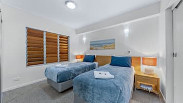 Quarto de hotel com duas camas, janela com persianas, mesa de cabeceira com abajur e um quadro na parede.