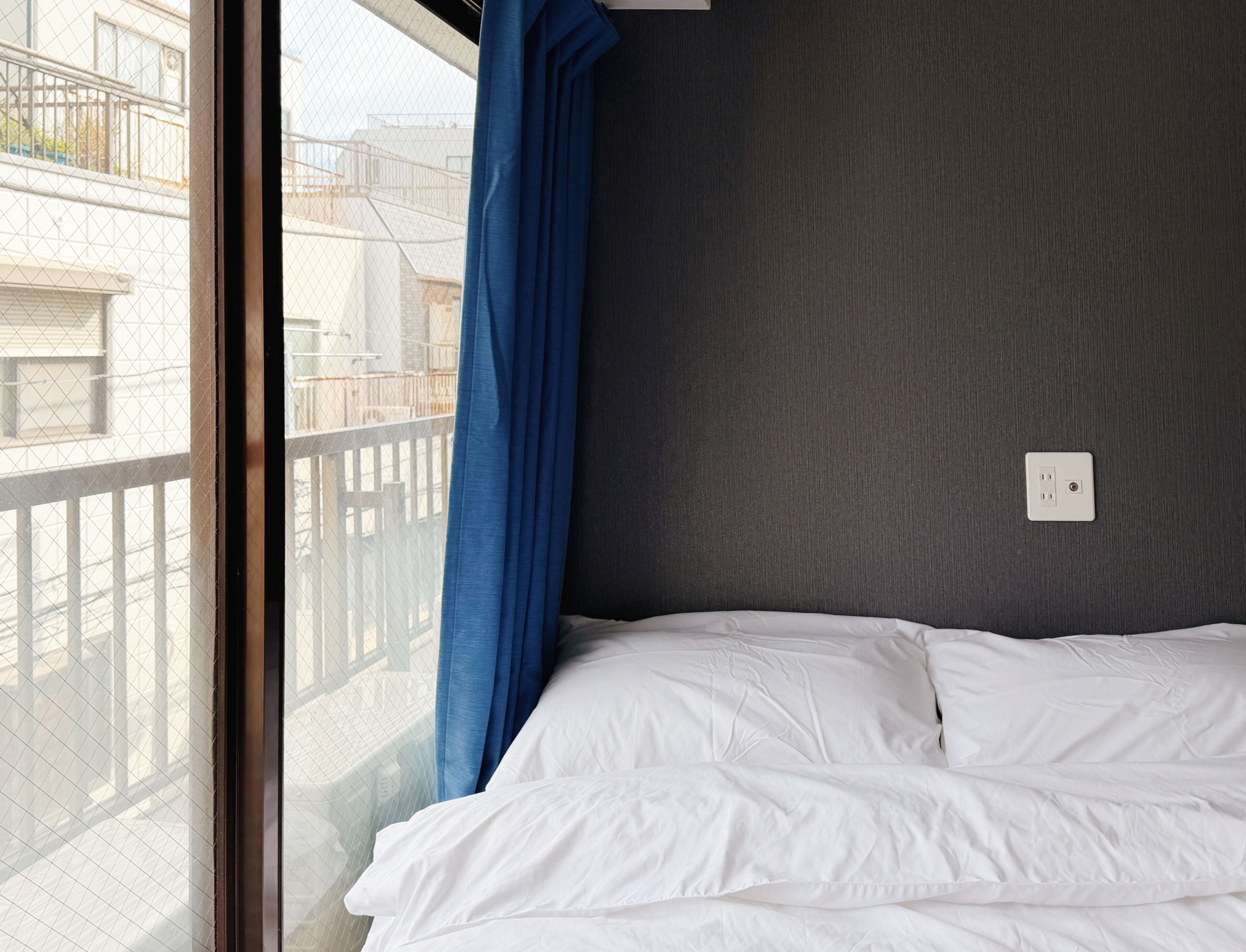 A bedroom with a bed, a window with a view of buildings, and a power outlet on the wall.