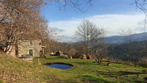 Ein rustikales Steinhaus mit einem kleinen Teich, umgeben von Bäumen und einer Bergkulisse.