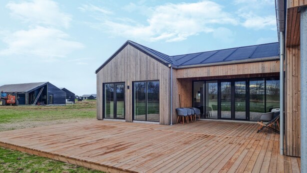Una casa moderna con una terrazza in legno, ampie porte scorrevoli in vetro e vista panoramica sul paesaggio circostante.