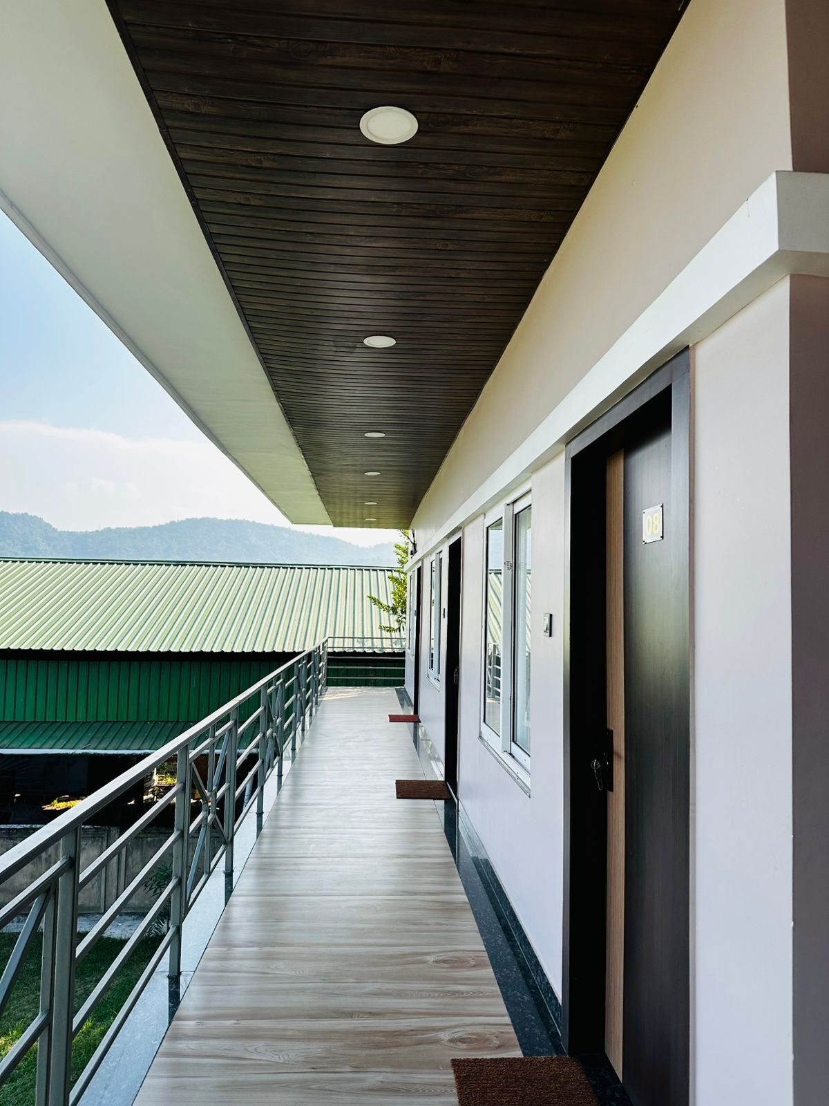 Un couloir doté d’un plafond en bois, d’une rambarde en métal et offrant une vue sur des bâtiments verdoyants et des montagnes au loin.