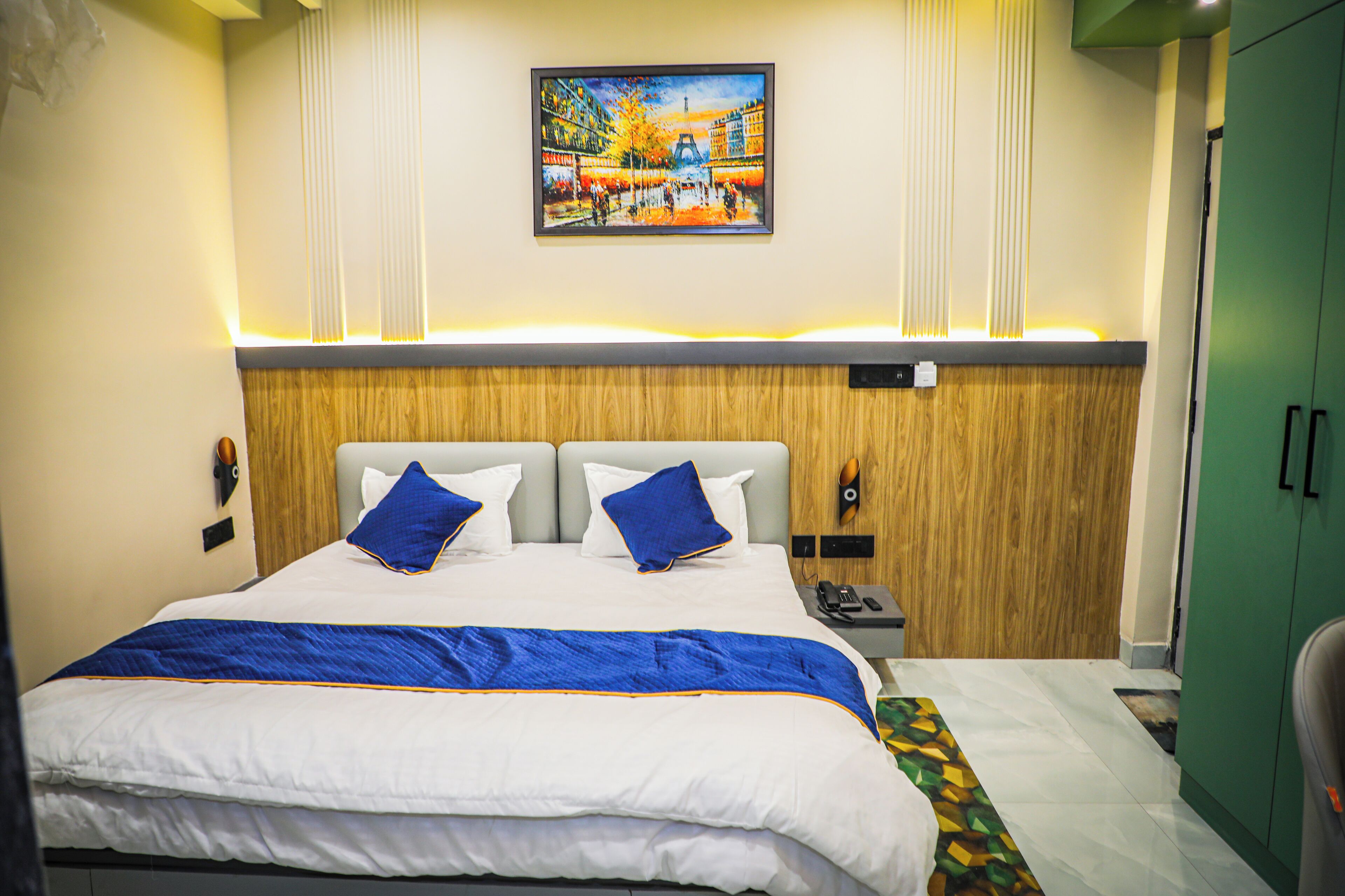A hotel room with a large bed, two pillows, a wooden headboard, a painting on the wall, and a green cabinet.