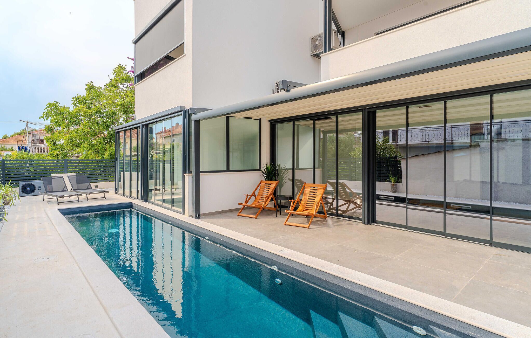 Un espace piscine extérieur moderne, avec une piscine à l’eau cristalline, deux chaises longues en bois et une terrasse fermée par des baies vitrées.