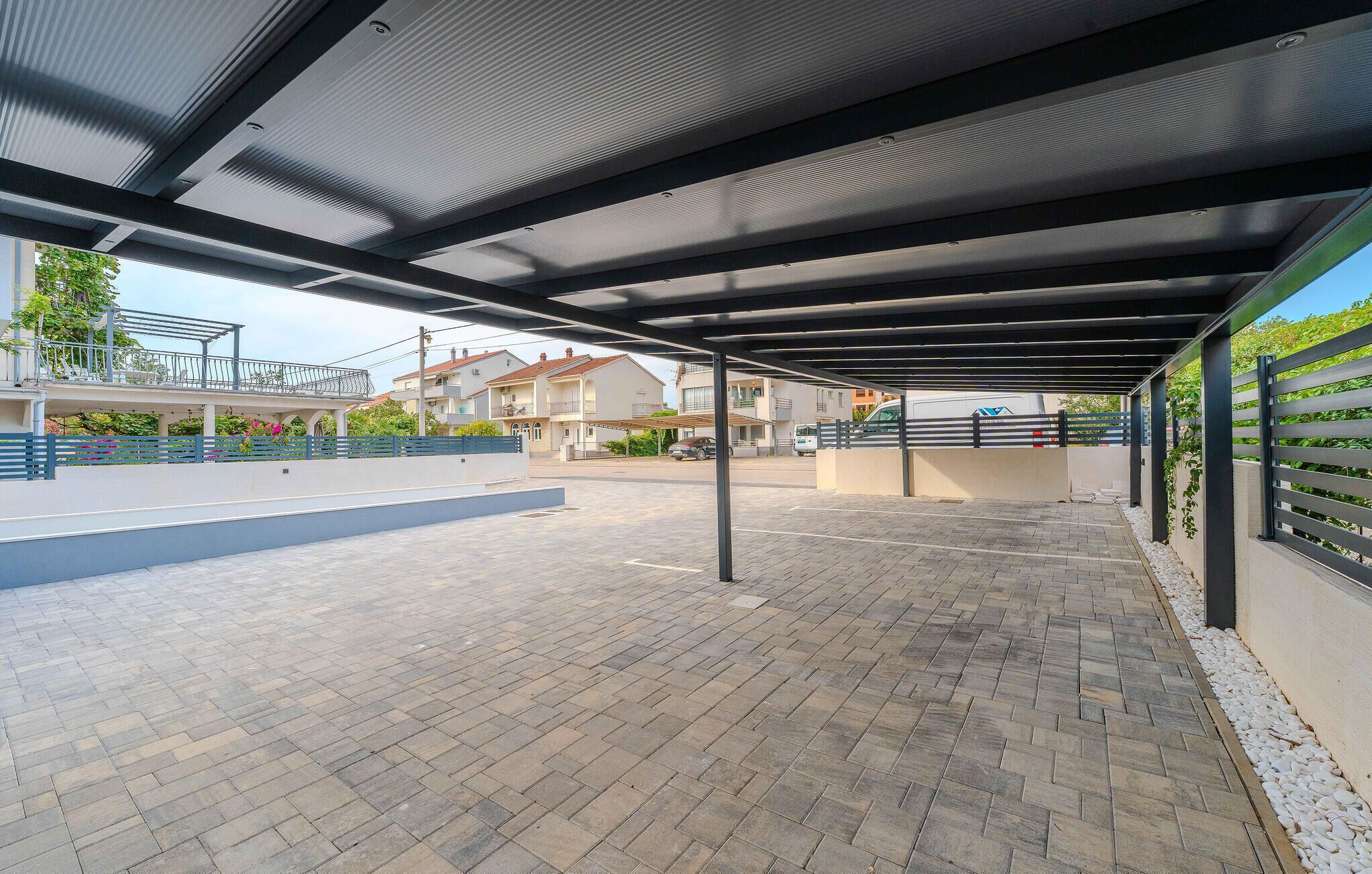 Ein überdachter Außenparkplatz mit befestigtem Weg, Metallgeländern und Blick auf Wohnhäuser im Hintergrund.