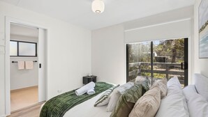 A modern bedroom with a bed, a small bedside table, a window with a view of the outdoors, and a door leading to another room.