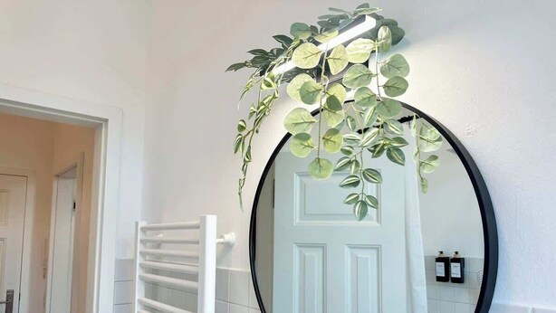 Une salle de bain avec un miroir rond, des murs recouverts de carreaux blancs, un porte-serviettes blanc et une porte.