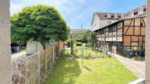 Une cour avec un grand arbre, une clôture en bois et un bâtiment dont la maçonnerie est apparente et qui est doté de lucarnes.
