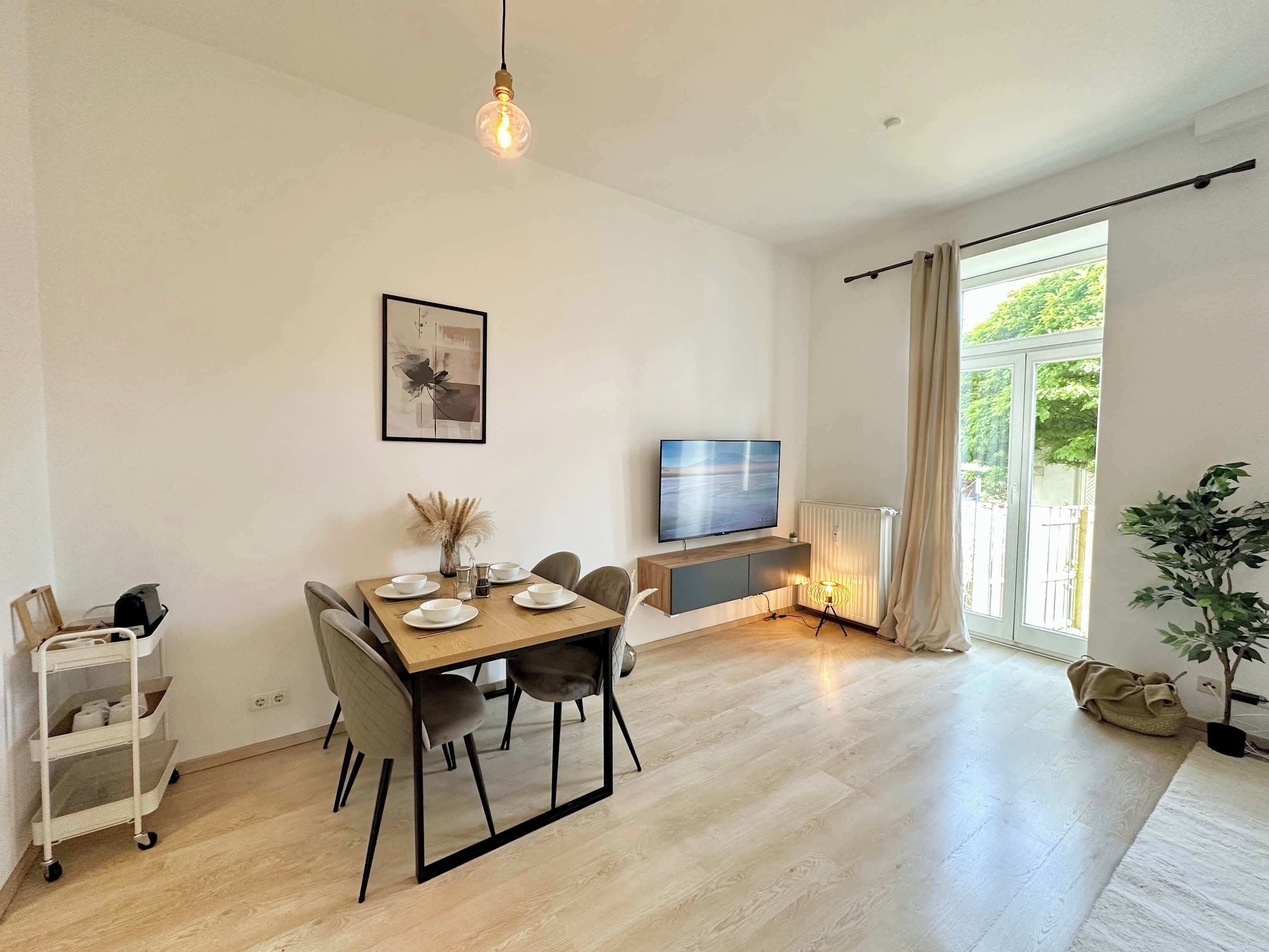 A modern dining area with a wooden table set for four, a television on a stand, and a large window with curtains.