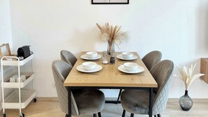 Un coin repas avec une table en bois dressée pour quatre personnes, entourée de chaises rembourrées. Une étagère blanche, sur laquelle se trouvent une machine à café et divers accessoires, est placée à gauche. Un tableau abstrait encadré est accroché au mur, au-dessus de la table.