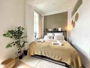 Een slaapkamer met een bed, een potplant, een nachtkastje en een raam.