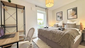 A bedroom with a bed, a desk, a chair, a hanging light, and a window.