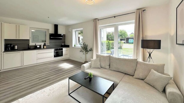 A modern living room with a sofa, coffee table, and a TV. There is a kitchen area with cabinets and a window.