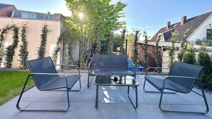 Eine Terrassengruppe im Freien mit drei schwarzen Metallstühlen und einem kleinen Tisch, umgeben von einem Betonboden und einem Metallzaun.