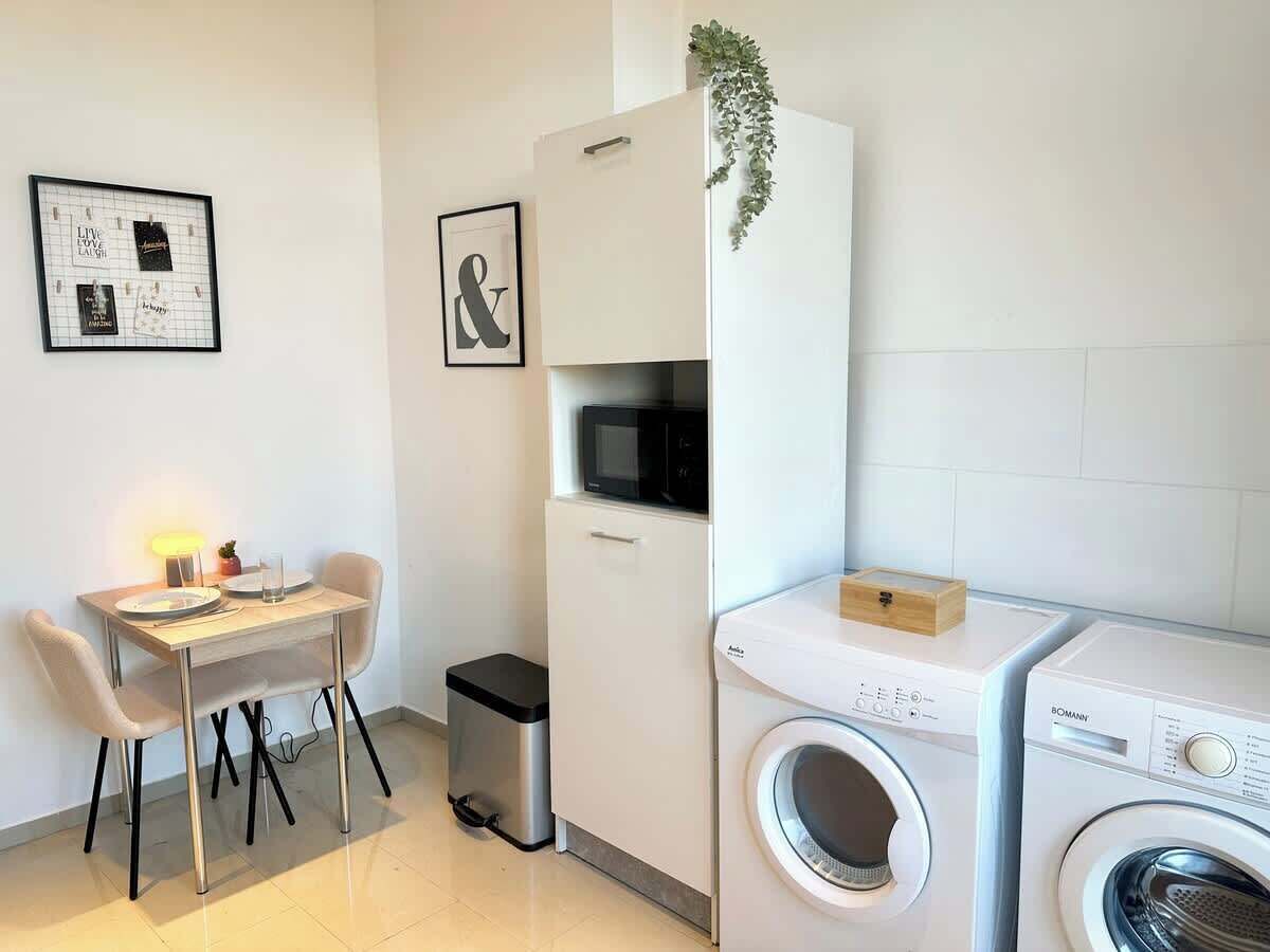 A compact kitchen with a microwave, a washing machine, and a small dining table with chairs.