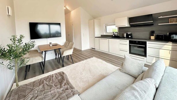 Una sala de estar moderna con un sofá, una mesa de centro y un televisor. Hay una zona de cocina con armarios y electrodomésticos.