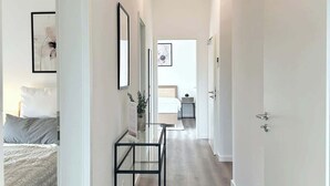 Un pasillo moderno y bien iluminado con una cama a la izquierda, una mesita y una puerta a la derecha.