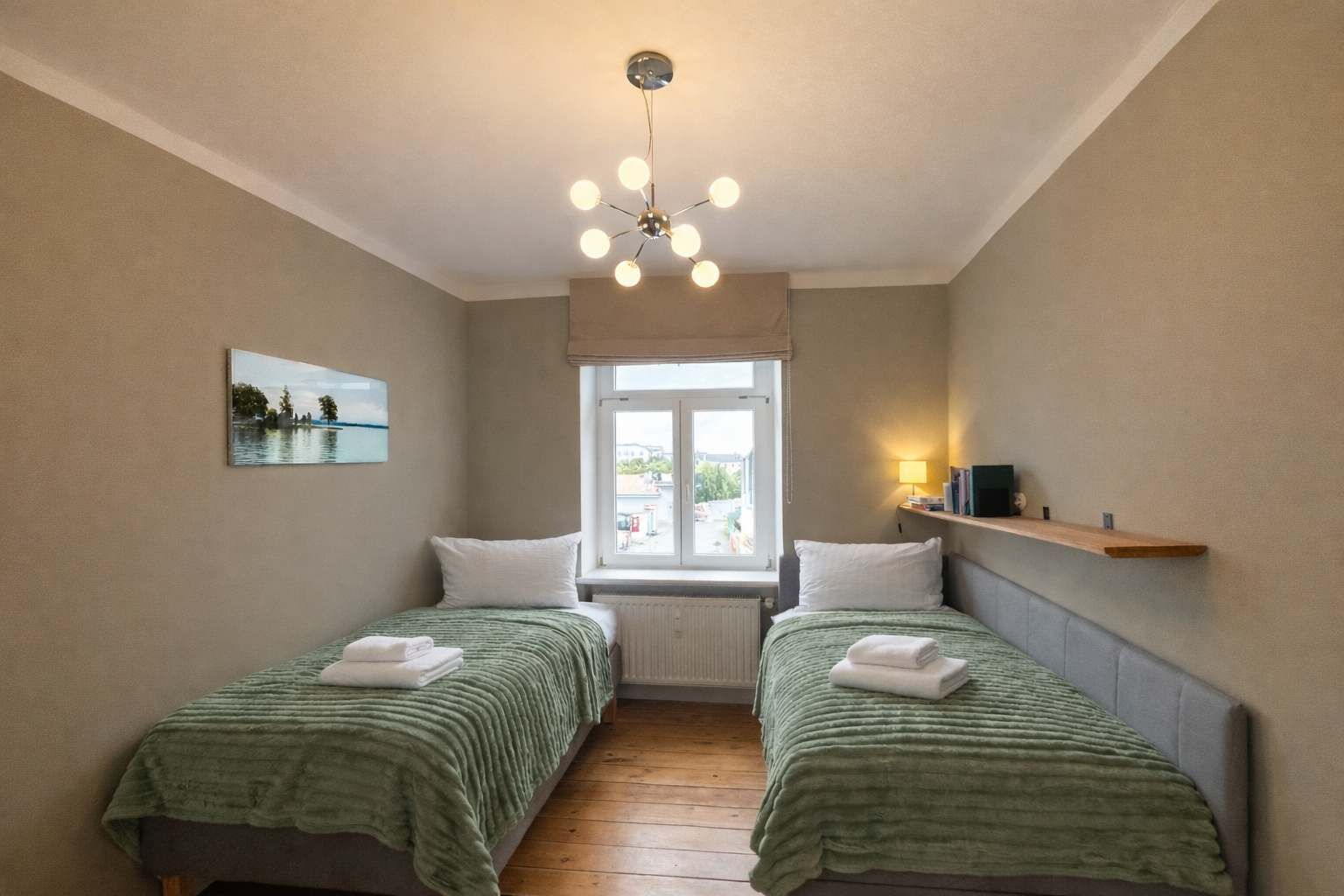 Ett litet, väl upplyst sovrum med två enkelsängar, ett fönster med utsikt, en taklampa och en hylla med böcker och en lampa.