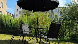 Eine Terrasse im Freien mit Tisch und Stühlen, einem Sonnenschirm und Blick auf Gebäude und Grünflächen.
