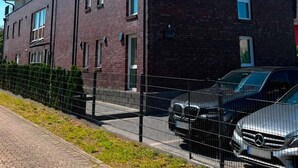 Ein mehrstöckiges Backsteinhaus mit flachem Dach, einem Balkon im oberen Geschoss und einem eingezäunten Parkplatz mit Autos.