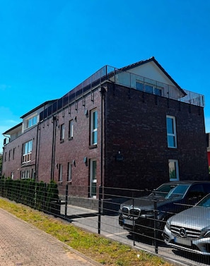 Un immeuble à plusieurs étages, construit en briques, avec un toit plat, un balcon à l’étage supérieur et un parking clos où se trouvent des voitures.