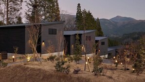 A modern house with a dark exterior, surrounded by a landscaped garden with small trees and shrubs, set against a backdrop of mountains. - Rakuten STAY VILLA Yufuin Onsen (Yufu)