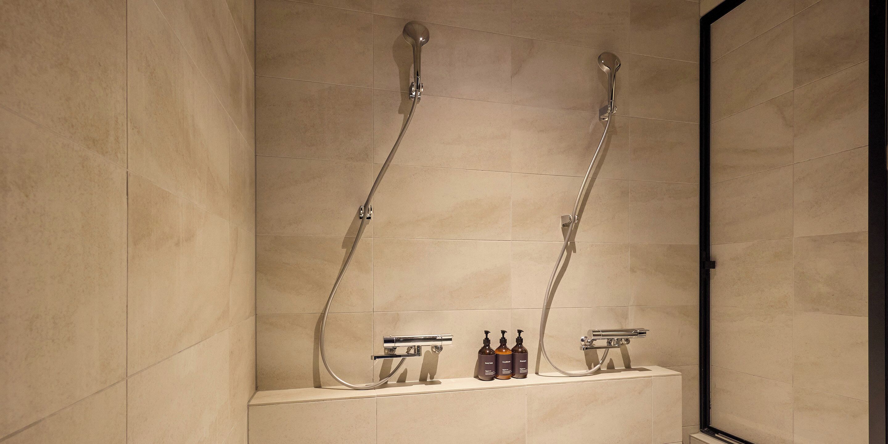 A bathroom with two showerheads, a sink, and three bottles of soap on a shelf.