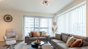 A modern living room with a sofa, armchair, and coffee table, large windows with blinds, and a city view. - Cosy apartment for 6 people with A/C, WIFI, TV and balcony (Bruges)