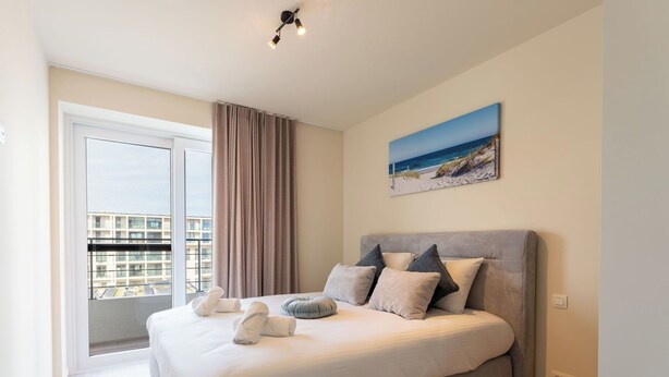 A modern bedroom with a large bed, a view of the city, and a painting of a beach scene. - Cosy apartment for 6 people with A/C, WIFI, TV and balcony (Bruges)