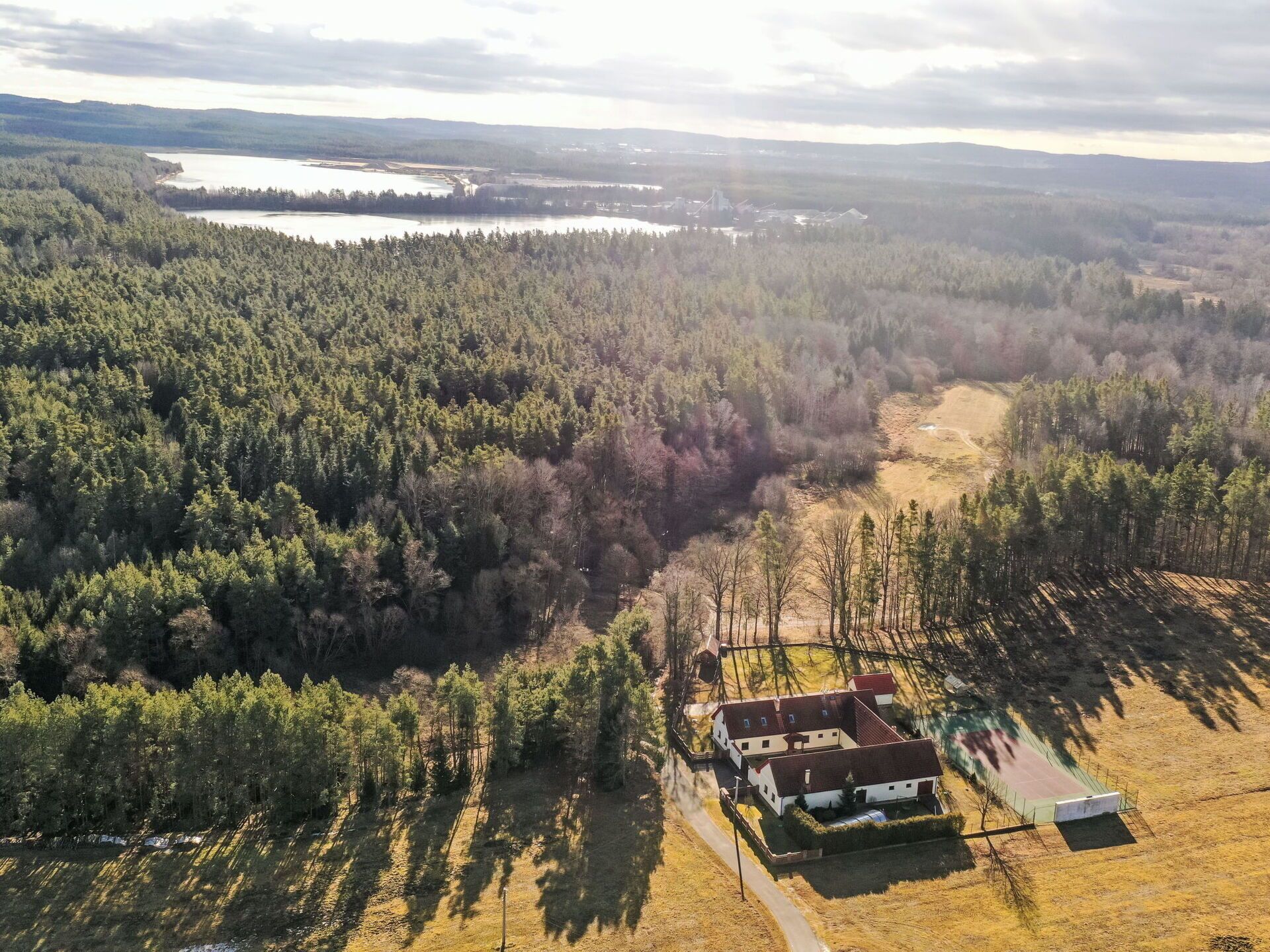 An aerial view of a property with a house, a fenced sports field, and surrounding forested area.