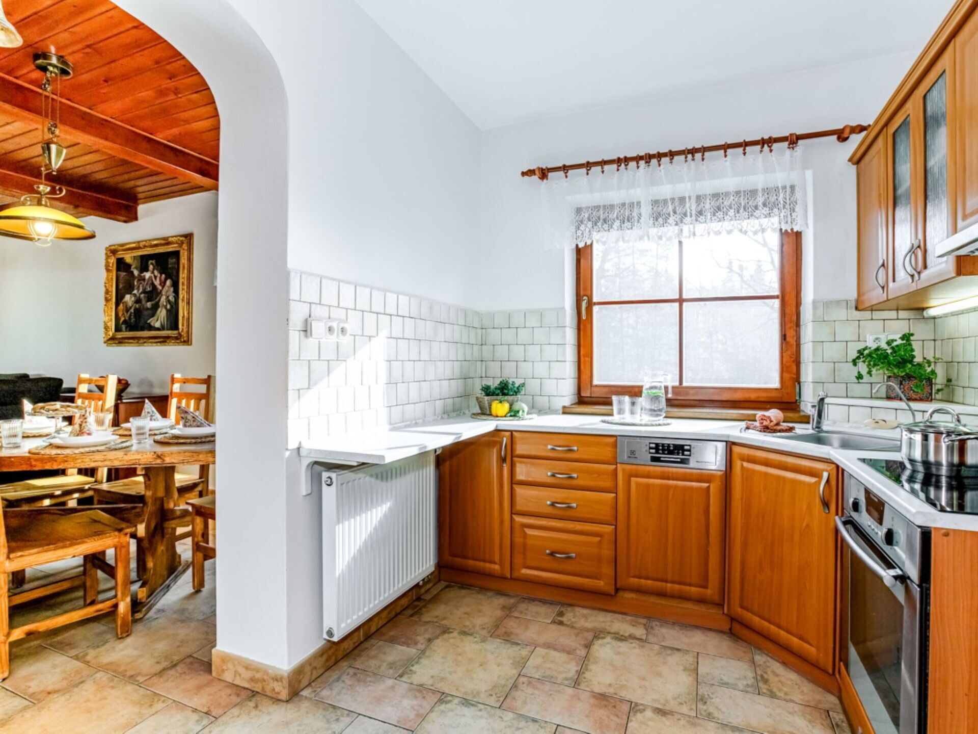 A kitchen with wooden cabinets, a white tiled backsplash, a window with lace curtains, and a dining area with a table and chairs.