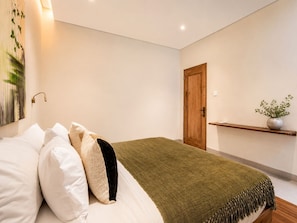 Ein modernes Schlafzimmer mit Bett, Kissen, einer grünen Tagesdecke, einer Holztür, einem an der Wand befestigten Regal mit Pflanze und einem Gemälde.