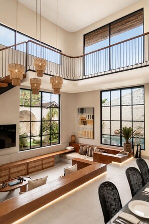 Un vaste espace de vie doté d’un haut plafond, d’une banquette intégrée, d’un coin repas avec des chaises et de grandes fenêtres offrant une vue sur l’extérieur.