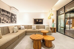 Un salon moderne avec un canapé d’angle, deux petites tables en bois et un grand téléviseur à écran plat fixé au mur.