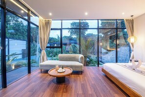 Une chambre d’hôtel moderne avec de grandes fenêtres, un canapé, un lit et une table ronde en bois.