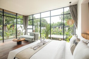 Une chambre d’hôtel moderne dotée d’un grand lit, d’un coin salon avec une table ronde et offrant une vue sur une végétation luxuriante grâce à de grandes fenêtres.