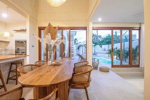 A dining area with a wooden table and wicker chairs, large windows with a view of a swimming pool and outdoor area. - 4BR Tropical Retreat With Private Pool Near Canggu (Canggu)