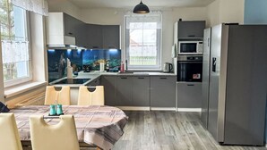 A modern kitchen with grey cabinets, a dining table with chairs, a large window with curtains, and a hanging pendant light.
