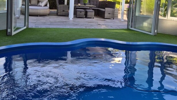 Un área de piscina moderna al aire libre con un porche de cristal, una pérgola blanca y mobiliario exterior.