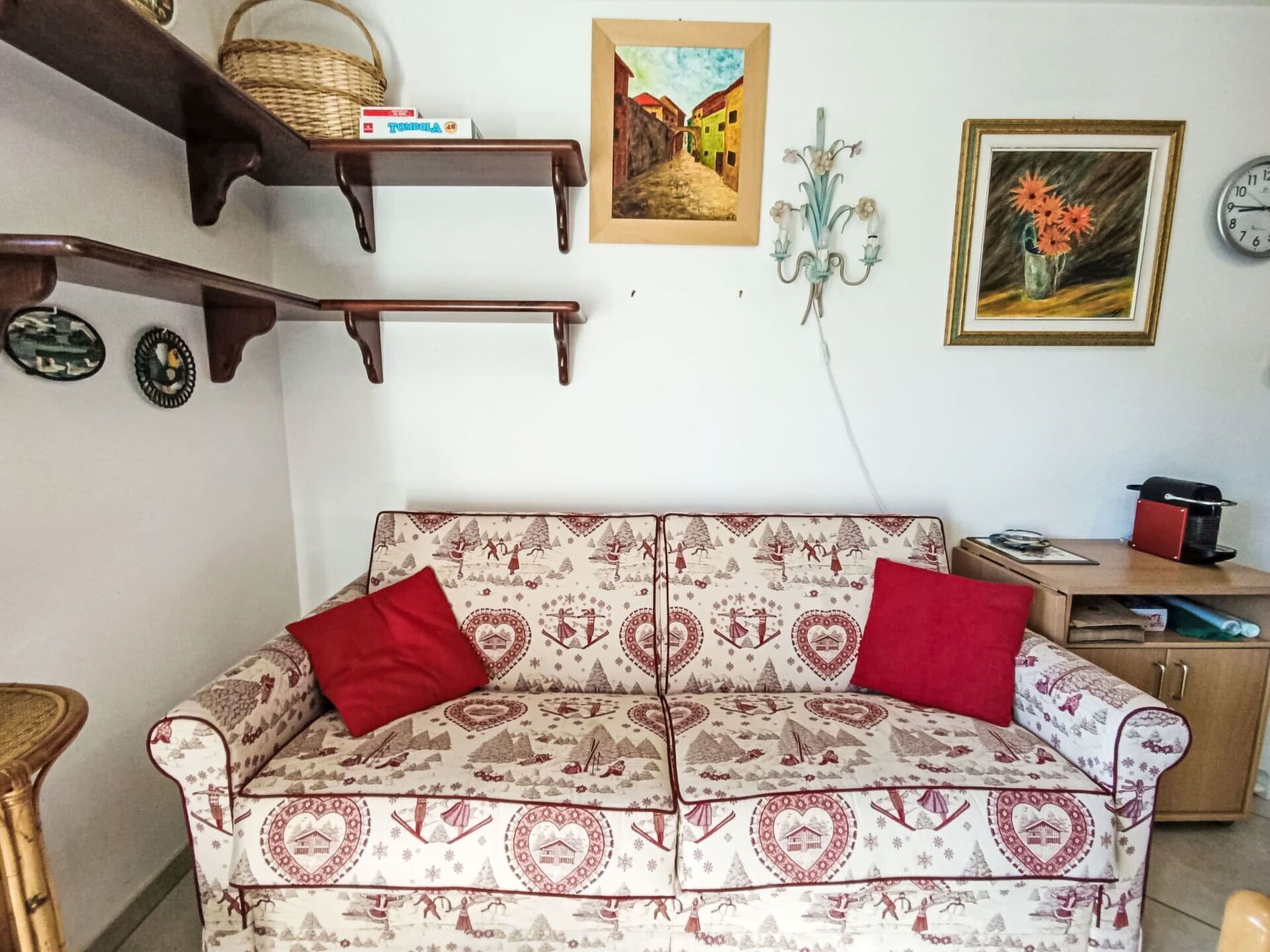 Ein gemütliches Wohnzimmer mit einem gemusterten Sofa, einem Holzregal mit einem Korb, einem gerahmten Gemälde, einer wandmontierten Kerzenleuchter und einer Uhr.