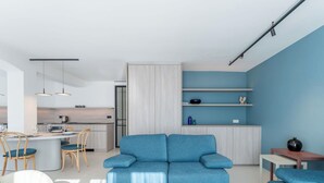 A modern living room with a blue sofa, a white armchair, and a wooden coffee table. There is a kitchen area with a dining table and chairs.