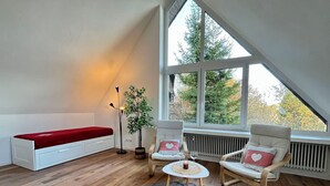Un acogedor dormitorio tipo loft con techo inclinado, un banco rojo, dos mecedoras, una mesita con una vela y un amplio ventanal con vistas a árboles.