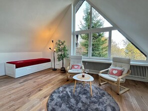 Ein gemütliches Schlafzimmer im Loft-Stil mit schrägem Dach, einer roten Bank, zwei Schaukelstühlen, einem kleinen Tisch mit Kerze und einem großen Fenster mit Blick auf Bäume.