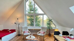 Un dormitorio estilo loft acogedor con techo inclinado, un ventanal con vistas a árboles, una cama con ropa de cama roja, dos sillones, una mesita con una vela y un comedor con mesa y sillas.