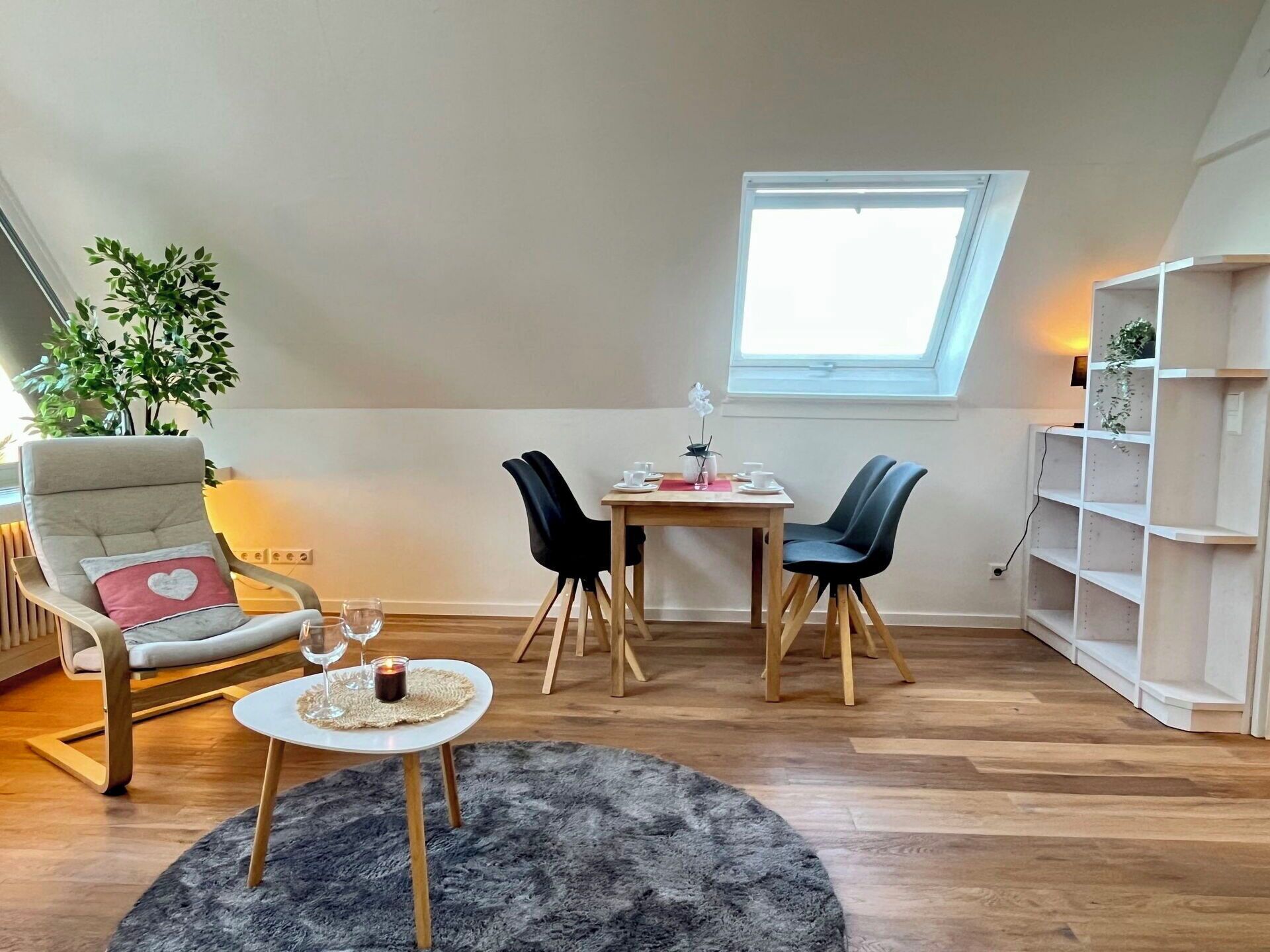 A cozy room with a high ceiling, a dining table with chairs, a small round table with a candle, a cushioned chair with a heart pillow, a potted plant, and a window with natural light.