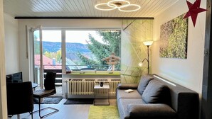 Un salon moderne avec un canapé, une table basse et un téléviseur. Une fenêtre offre une vue sur les arbres et un immeuble.