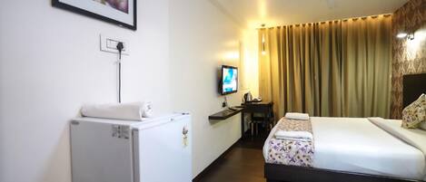 A hotel room with a bed, a desk with a chair, a television, a refrigerator, and a wall-mounted artwork.