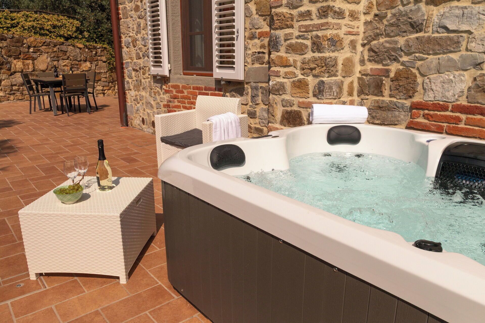 A hot tub with water, a bottle of wine, and glasses on a table, set against a stone wall with a wicker chair and a table in the background.