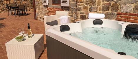 A hot tub with water, a bottle of wine, and glasses on a table, set against a stone wall with a wicker chair and a table in the background.